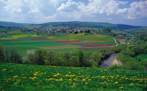 Zielone pogórze z kwitnącymi na żółto łąkami, polami uprawnymi i wijącą się rzeką. W oddali widać zabudowania i lasy.