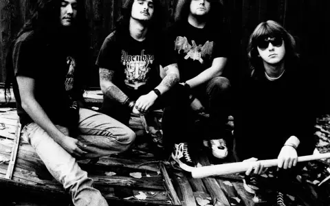 Vier Männer in Band-T-Shirts und Jeans sitzen auf Holz. Einer hält einen Baseballschläger.