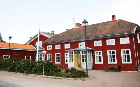 Röda trähus med vita fönster och tegeltak. En skylt visar "Stiftsgården Undersvik". Vackert beläget med natur och vatten i bakgrunden.
