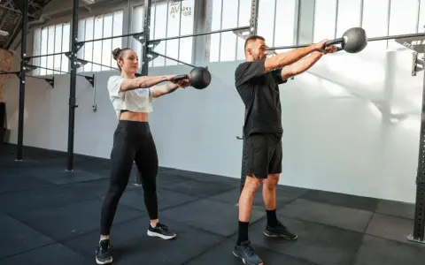 Kobieta i mężczyzna wykonują ćwiczenia z kettlebellami w siłowni.