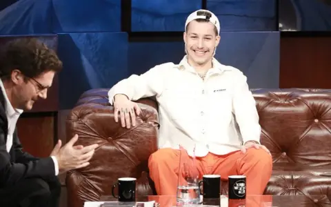 Zabson, z uśmiechem na twarzy, siedzi na kanapie w studiu. Obok niego siedzi mężczyzna w okularach.