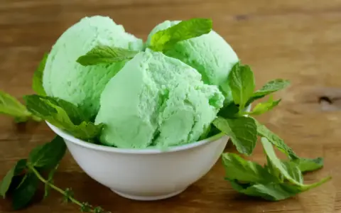 Trois boules de glace verte menthe, rappelant la saveur de la glace Get 27, garnies de feuilles de menthe fraîche dans un bol blanc sur fond de bois.