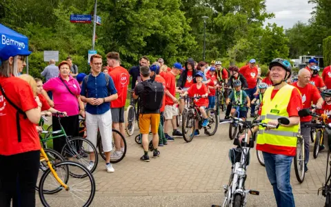 Uczestnicy rajdu rowerowego w czerwonych koszulkach, dzieci i dorośli, gotowi do startu.
