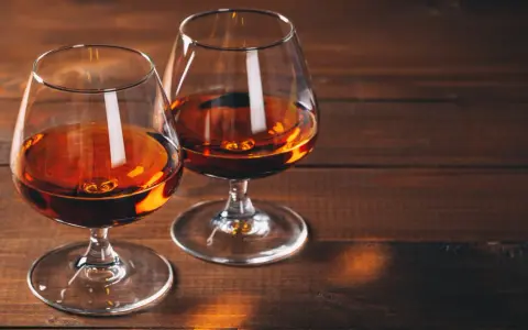 Dwa kieliszki z bursztynowym płynem, przypominającym bourbon lub whiskey, stoją na drewnianym stole.