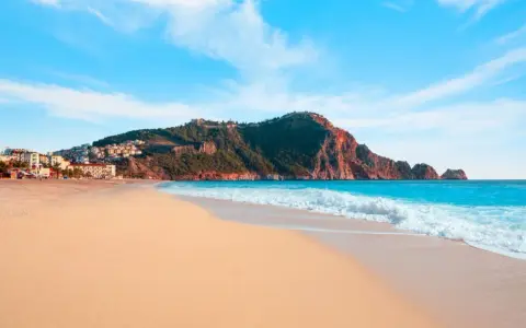 Plaża Kleopatry w Alanyi z turkusową wodą, piaszczystym brzegiem i górującym nad nią masywem skalnym z twierdzą.