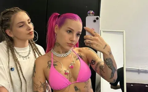 Dwie młode kobiety, jedna z różowymi włosami i tatuażami, druga z warkoczami, pozują do selfie.