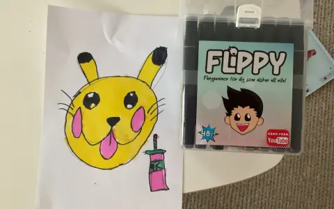 En gul Pikachu-liknande figur med rosa kinder och en tunga som sticker ut, bredvid en låda med färgpennor "FLIPPY" för dig som älskar att rita.