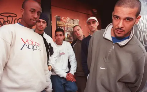 Un groupe de rap français des années 90/2000 pose devant un mur tagué. Ils portent des sweats et des jeans, typiques de l'époque.