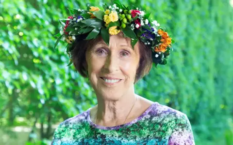 Eva Rydberg ler glatt med en blomsterkrans på huvudet, omgiven av grönska.