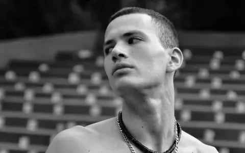 Joven con un corte de pelo buzz cut largo, mirando hacia un lado con una expresión pensativa. Lleva un collar de cadena.