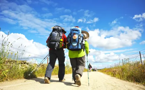 Två vandrare med ryggsäckar går längs en grusväg under en blå himmel med moln. En perfekt dag för pilgrimsvandring i Sverige.