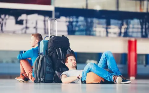 Dwóch mężczyzn z bagażami czeka na lotnisku. Jeden śpi, drugi czuwa. Zastanawiasz się, ile wcześnie być na lotnisku?