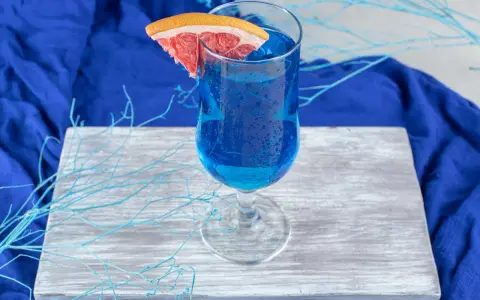 Przepis na Blue Lagoon: Błękitny drink z plasterkiem grejpfruta, ozdobiony niebieskimi gałązkami, na tle granatowej tkaniny.
