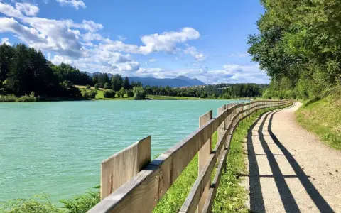 Sentiero sterrato con staccionata in legno lungo il lago di Tavon, circondato da alberi e montagne. Cielo azzurro con nuvole bianche.