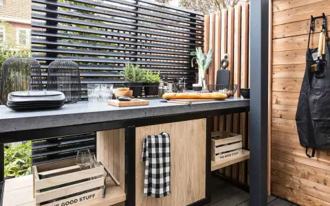Cucina all'aperto fai da te con piano in cemento, pane, verdure e utensili. Un grembiule nero è appeso a una parete di legno.