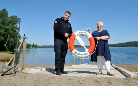 Två personer håller i en räddningsboj vid en sjö. En man i uniform och en kvinna ler. Trygg-Hansa vid Nörybyns camping.