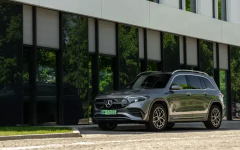 Nowy Mercedes 4x4, elegancki SUV, zaparkowany przed nowoczesnym budynkiem z zielonymi odbiciami drzew w szybach.