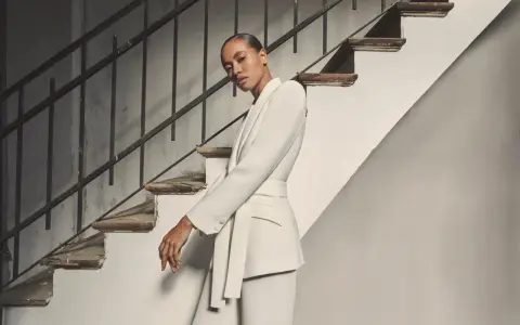 Mujer posando con un elegante outfit blazer blanca, pantalón a juego y cinturón, junto a una escalera antigua.