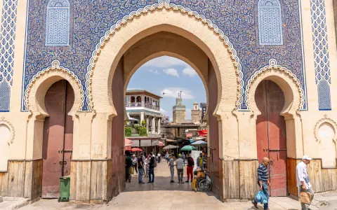 Prachtvolles Tor in Fes Stadt mit blau-goldenen Mustern. Menschen beleben den Platz dahinter.