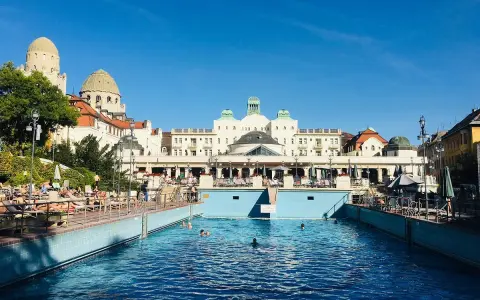 Soligt utomhuspool vid ett termalbad i Budapest. Människor solar och badar i det blå vattnet framför en imponerande byggnad.