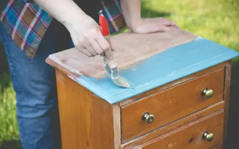 Persona pinta con brocha azul una mesita de noche de madera, transformándola en una de las mesitas de noche pintadas a la tiza.
