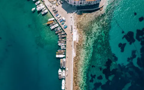 Widok z lotu ptaka na port z licznymi łodziami i jachtami cumującymi przy nabrzeżu. Woda jest krystalicznie czysta, a na dnie widać kamienie i ciemne plamy wodorostów.