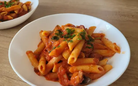 Penne z sosem pomidorowym i kawałkami kiełbasy, posypane natką pietruszki. Pyszny makaron z kiełbasą na białym talerzu.