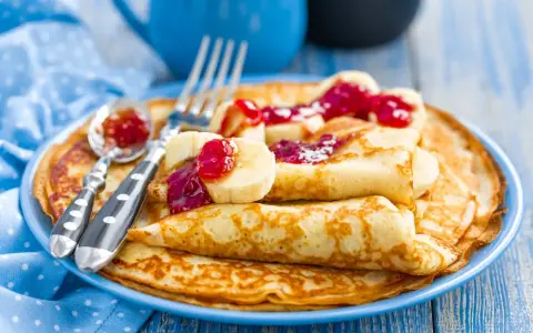 Stos idealnie przygotowanego ciasta na naleśniki, polanego dżemem i kawałkami banana. Obok leżą sztućce.