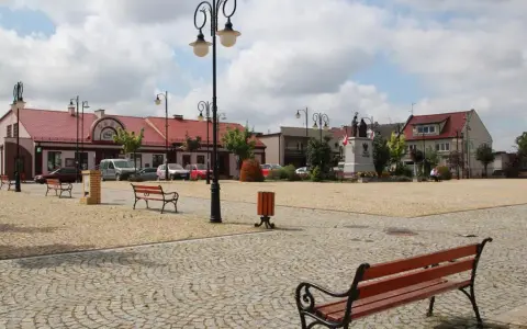 Rynek w Ślesinie z pomnikiem, ławkami i budynkami. Atrakcje tego miejsca zachęcają do odpoczynku.