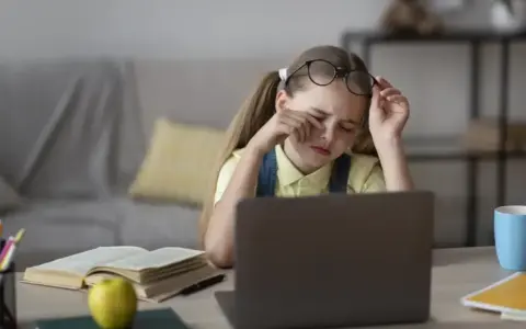 Zmęczona dziewczynka z okularami na czole pociera oczy, ucząc się przy laptopie. Jej indywidualna ścieżka nauczania jest trudna.