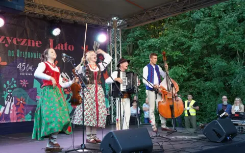 Zespół w strojach ludowych gra na scenie. Może to być część koncertu z piosenkami o Polsce dla dzieci.