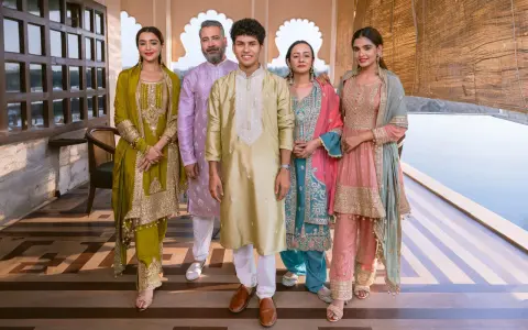 A family poses in vibrant traditional clothing from India. The women wear ornate salwar kameez sets in olive green, teal, and pink, while the men are dressed in elegant kurtas.