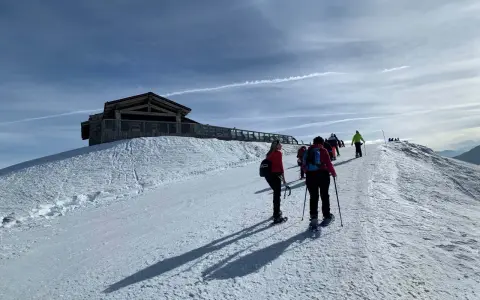 Gruppo di escursionisti con ciaspole su un sentiero innevato, con un rifugio in legno in cima a una collina. Posti belli in montagna Piemonte inverno.
