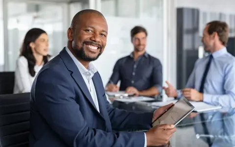 The executive board chair smiles, holding a tablet in a meeting with colleagues.