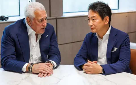 Koji Watanabe conversa con un hombre mayor, ambos con trajes azules. Un modelo de coche deportivo negro se ve en el fondo.