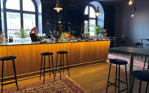 En bar med träpanel och svarta barstolar. En person bakom baren förbereder en drink. En meny på svarta tavlan visar priser för drycker i det musikaliska kvarteret.