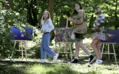 Trzy dziewczyny spacerują wśród obrazów na sztalugach. Jedna z nich, w niebieskich jeansach, przypomina młodszą wersję Tomasza Kłosowskiego.