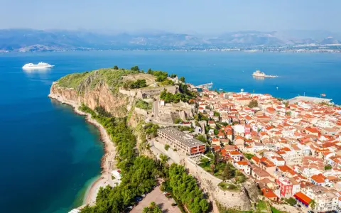 Widok na Nafplio w Grecji: miasto z czerwonymi dachami, twierdza na skale i biały statek wycieczkowy na lazurowym morzu.
