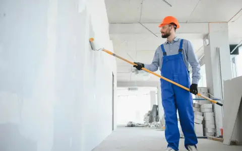 Maler mit blauer Latzhose und orangefarbenem Helm streicht eine Wand weiß. Informieren Sie sich über die malerarbeiten kosten für Ihr Projekt.