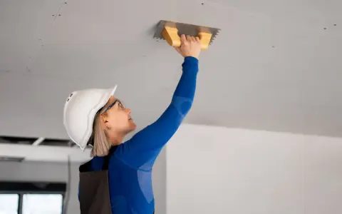 Kobieta w kasku i okularach wykonuje tynkowanie sufitu przy użyciu kielni.
