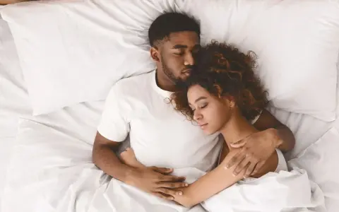Ein Paar schläft eng zusammen in einem Bett. Er hält sie schützend im Arm, während sie sich an ihn kuschelt.
