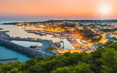 Porto illuminato al tramonto, un invito al turismo Salento. Barche ormeggiate e case che brillano sulla costa.