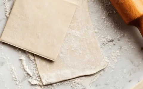 Pâte feuilletée prête à être travaillée, saupoudrée de farine sur un plan de travail en marbre. Un rouleau à pâtisserie en bois est visible sur la droite.