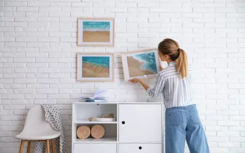 Mujer cuelga cuadros de paisajes marinos en pared de ladrillo blanco. Aprende como colocar cuadros de diferentes tamaños para un diseño armonioso.