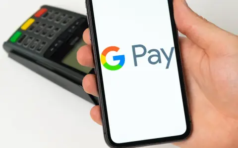 Dłoń trzyma smartfon z widocznym logo Google Pay. W tle terminal płatniczy. Szybkie i wygodne płatności.