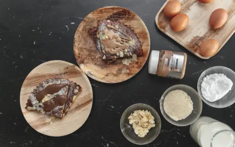 Pyszny keto omlet na słodko z kremem migdałowym, płatkami migdałów i cukrem pudrem. Obok składniki: jajka, mleko, mąka migdałowa, erytrytol.