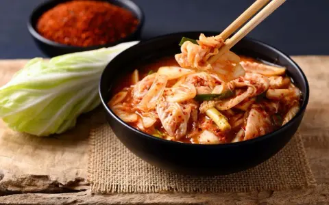 Miseczka z kimchi, pałeczki podnoszące kawałek kapusty, obok kapusta i miseczka z przyprawą.