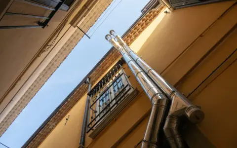 Dos tubos de salida humos estufa pellets brillan bajo el sol, ascendiendo por la pared amarilla de un edificio.