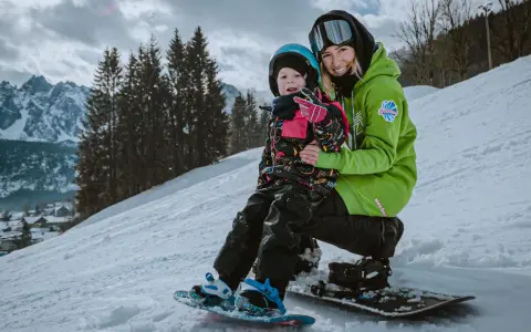 Uśmiechnięta instruktorka na stoku narciarskim dla dzieci trzyma małego snowboardzistę.