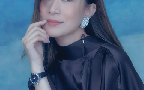 A woman with a thoughtful expression, her finger near her lips, wears a dark satin dress, a striking earring, and a blue-faced watch. She looks xa thi man.
